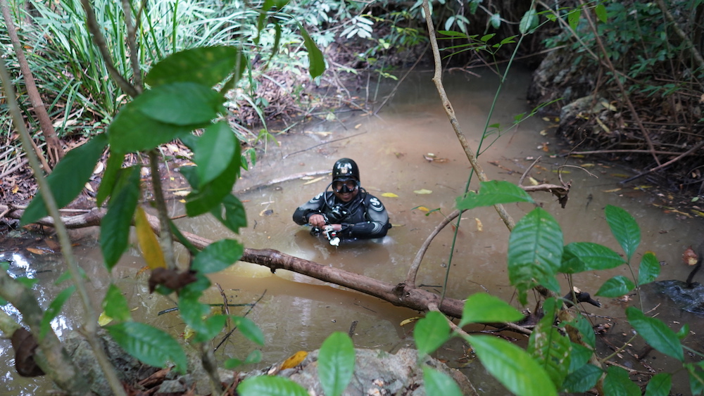 A sidemount diver standing chest-deep in a small, silty jungle pond at the Sra Keow cave entrance, Krabi
